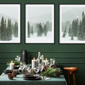May include: Three framed black and white photographs of a snowy forest scene with pine trees. The photos are hanging on a dark green wall above a table set for a holiday dinner. The table is covered in a green tablecloth and decorated with pine branches, pine cones, candles, and wine glasses.