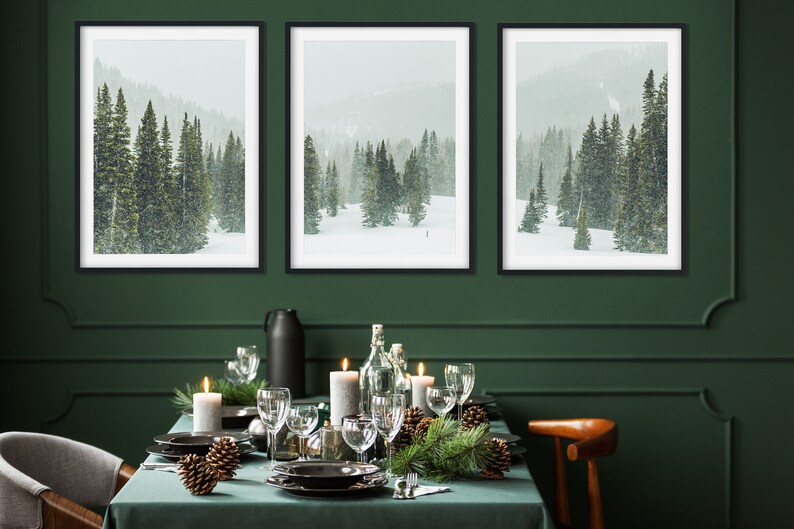 May include: Three framed black and white photographs of a snowy forest scene with pine trees. The photos are hanging on a dark green wall above a table set for a holiday dinner. The table is covered in a green tablecloth and decorated with pine branches, pine cones, candles, and wine glasses.