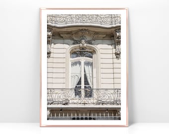 May include: A vintage-style window with ornate ironwork and a balcony. The window is framed by a white building with a cream-colored facade. The window has a white curtain and a view of a building across the street.