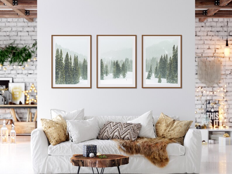 May include: Three framed prints of a snowy forest scene with tall evergreen trees. The prints are hung on a white wall above a white couch with throw pillows and a fur throw.