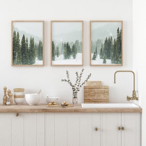 May include: Three framed prints of snowy forest scenes with evergreen trees. The prints are hung on a white wall above a wooden countertop with a white sink and gold faucet.