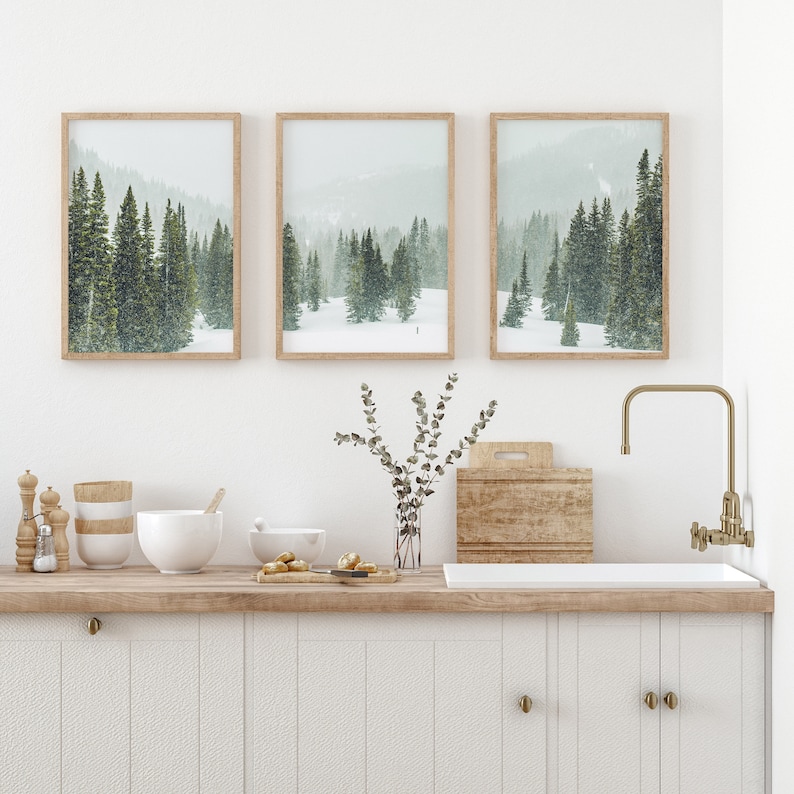 May include: Three framed prints of snowy forest scenes with evergreen trees. The prints are hung on a white wall above a wooden countertop with a white sink and gold faucet.