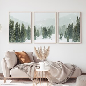May include: Three framed landscape photographs of a snowy forest with evergreen trees. The photos are hung on a white wall above a light beige couch with a brown throw pillow and a beige throw blanket. A vase with dried pampas grass sits on a gold metal side table.