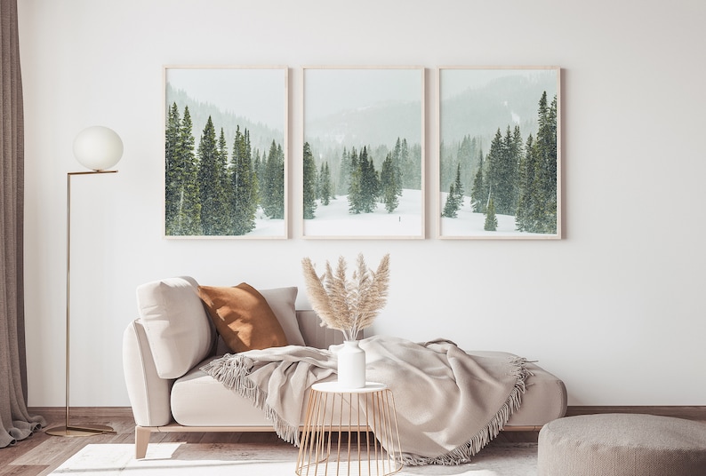 May include: Three framed landscape photographs of a snowy forest with evergreen trees. The photos are hung on a white wall above a light beige couch with a brown throw pillow and a beige throw blanket. A vase with dried pampas grass sits on a gold metal side table.