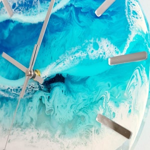May include: Round wall clock with a resin ocean design. The clock face features shades of blue and white, with silver-coloured hour markers and hands. The clock's design evokes a coastal theme.