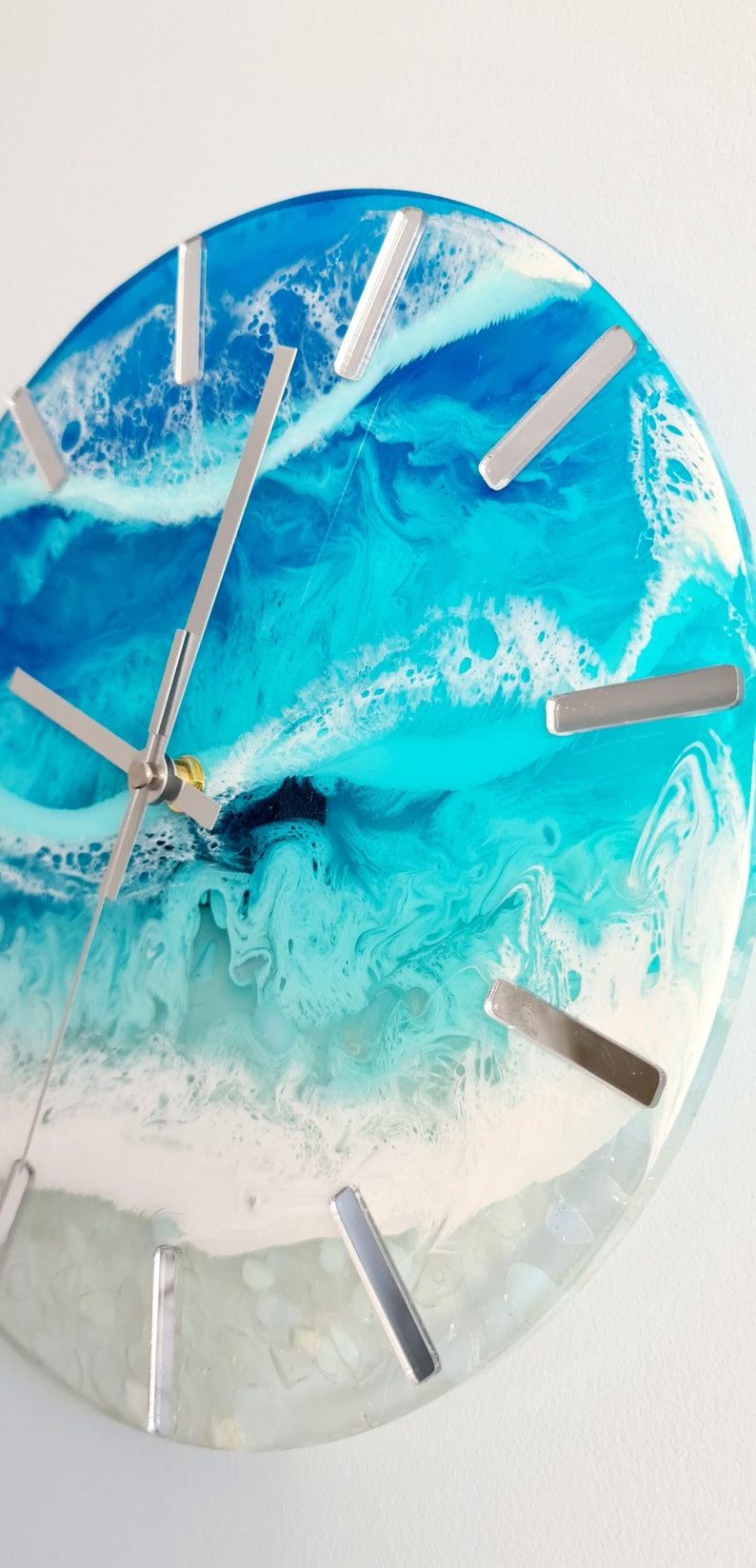 May include: Round wall clock with a resin ocean design. The clock face features shades of blue and white, with silver-coloured hour markers and hands. The clock's design evokes a coastal theme.