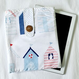 May include: A white tablet case decorated with beach hut appliqués in blue, red, and white. The case has a wooden button closure and is designed to fit a tablet. The huts have flags.