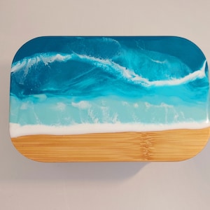 May include: A rectangular bamboo chopping board with a resin ocean design. The top half features layers of blue and white resin, mimicking ocean waves. The bottom half is natural bamboo, resembling a beach.