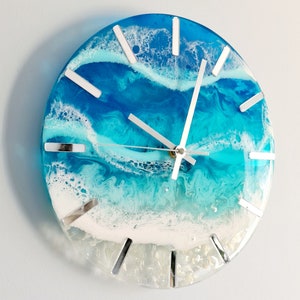 May include: Round wall clock with a resin ocean design. The clock face features shades of blue and white, with silver-coloured hands and hour markers. The clock measures approximately 25 cm in diameter.