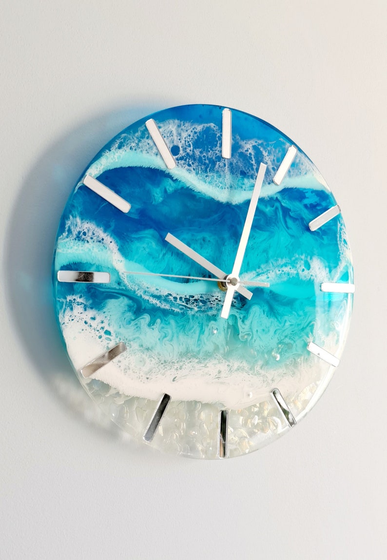 May include: Round wall clock with a resin ocean design. The clock face features shades of blue and white, with silver-coloured hands and hour markers. The clock measures approximately 25 cm in diameter.