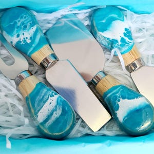 May include: A cheese knife set with blue and white resin handles and stainless steel blades. The handles have a wood base and silver accents. The set includes a cheese fork, a spreader, and various cheese knives, all presented in a white box with blue tissue paper.