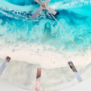 May include: A half-circle wall clock with a resin design resembling an ocean scene. The clock face features shades of blue, white, and grey, with silver-coloured hour markers and clock hands. The overall design evokes a coastal theme.