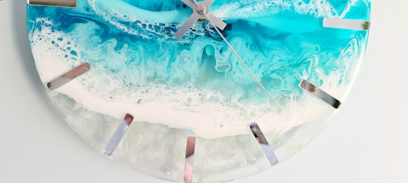May include: A half-circle wall clock with a resin design resembling an ocean scene. The clock face features shades of blue, white, and grey, with silver-coloured hour markers and clock hands. The overall design evokes a coastal theme.