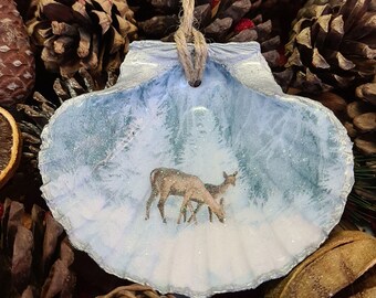 May include: A painted seashell Christmas ornament depicting a winter forest scene with two deer. The shell is painted with a light blue and white background, and has a silver glitter trim. A natural twine loop is attached for hanging.