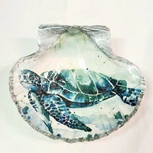 May include: A decorative seashell featuring a painted sea turtle design. The turtle is rendered in shades of blue and green, set against a watercolour-style background. The shell's edge is trimmed with a silver metallic finish, adding a touch of elegance.