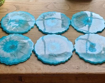 May include: Six resin coasters with an irregular, organic shape. Each coaster has a vibrant turquoise border and a central design with shades of blue and white, accented with glitter. These decorative coasters are perfect for protecting surfaces.