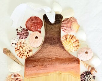 May include: A wooden chopping board with a resin design in shades of brown and peach, reminiscent of a beach. The board is surrounded by various seashells, including scallop shells and sea urchins, and a white ribbon.