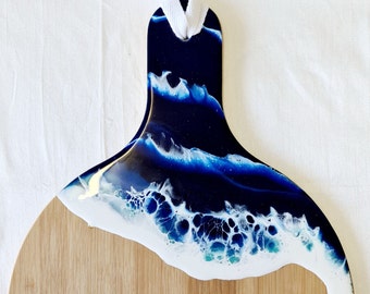 May include: A wooden serving board with a handle, featuring a resin design that mimics ocean waves. The resin art transitions from deep navy to white, creating a coastal aesthetic. A white ribbon is attached for hanging.