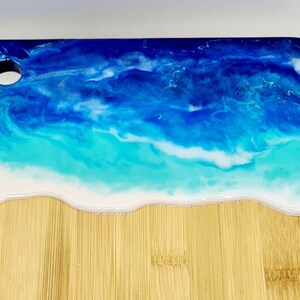 May include: A rectangular bamboo chopping board with a resin design. The top of the board features a blue and white resin design resembling ocean waves. The bottom portion of the board is natural bamboo.