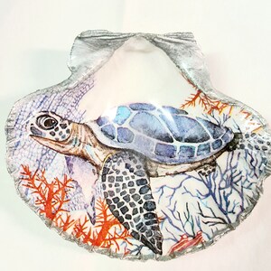 May include: A painted seashell with a sea turtle design. The turtle has a blue and white shell, with a detailed head and flippers. Coral and other sea life are depicted in shades of orange and blue.