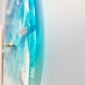 May include: A circular wall clock with a blue and white ocean design. The clock face has a swirling pattern of blue and white resin, with silver markers for the hours. The clock is mounted on a white wall.