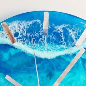 May include: A round wall clock with a resin ocean design. The clock face features shades of blue and white, with silver-coloured hour markers and hands. The clock's design evokes a coastal theme.