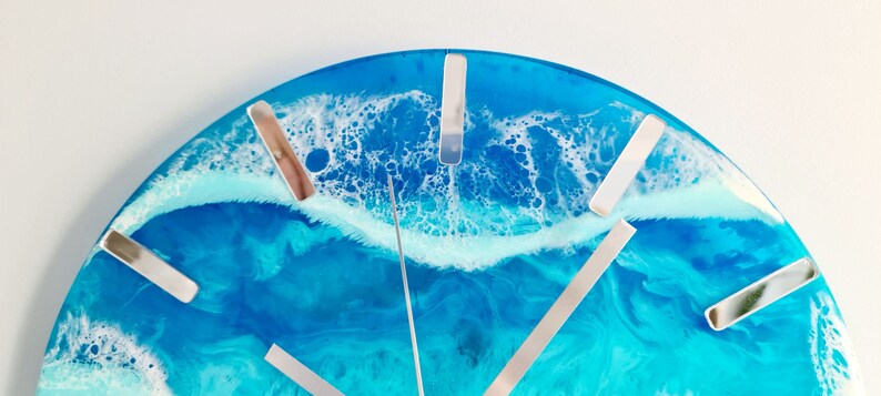 May include: A round wall clock with a resin ocean design. The clock face features shades of blue and white, with silver-coloured hour markers and hands. The clock's design evokes a coastal theme.
