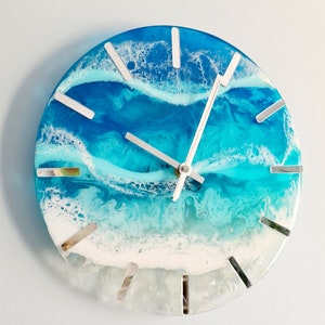 May include: Round wall clock with a resin ocean design. The clock face features shades of blue and white, mimicking ocean waves. Silver markers indicate the hours. The clock hands are silver.