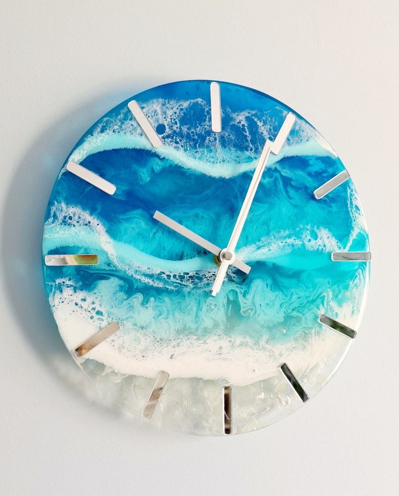 May include: Round wall clock with a resin ocean design. The clock face features shades of blue and white, mimicking ocean waves. Silver markers indicate the hours. The clock hands are silver.