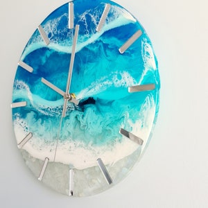 May include: Round wall clock with a resin ocean design. The clock face features shades of blue and white, with silver-coloured hour markers and clock hands. The clock has a coastal theme.