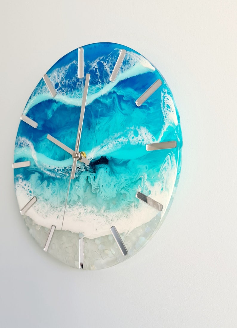 May include: Round wall clock with a resin ocean design. The clock face features shades of blue and white, with silver-coloured hour markers and clock hands. The clock has a coastal theme.