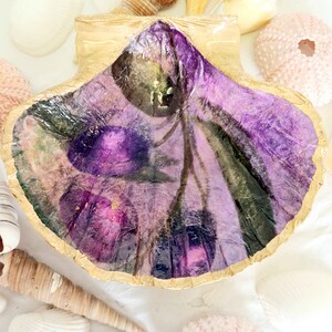 May include: A decorative seashell with a gold border and a purple floral design. Various seashells and a pink sea urchin surround the shell on a white surface. The shell's interior has a watercolour-style floral pattern. The shell is a beautiful decorative item.