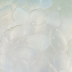 May include: Close-up of a clear glass bowl filled with smooth, pale blue-green stones. The stones have a rounded shape and a slightly translucent appearance. The bowl is set against a white background.