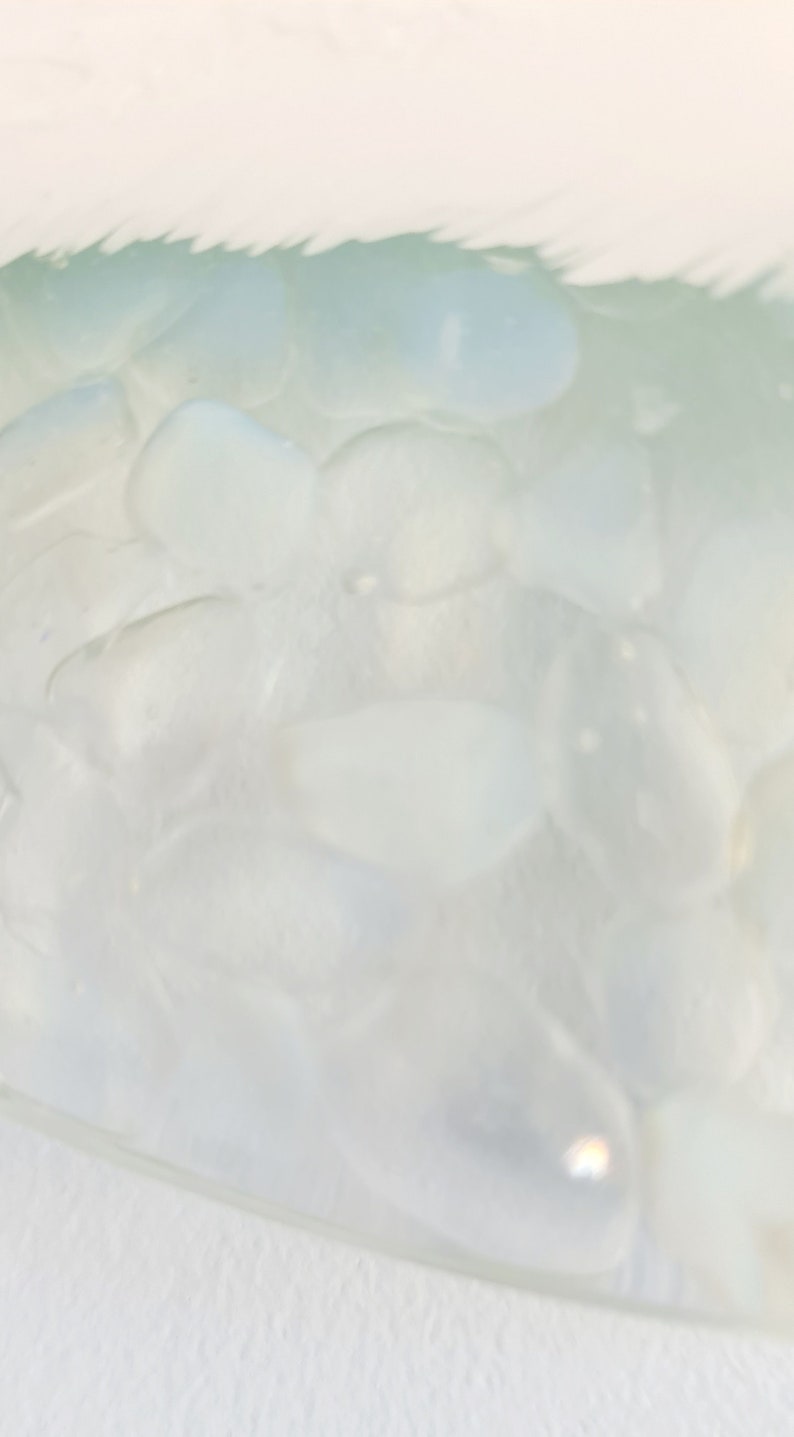 May include: Close-up of a clear glass bowl filled with smooth, pale blue-green stones. The stones have a rounded shape and a slightly translucent appearance. The bowl is set against a white background.