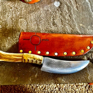 Könnte beinhalten: Ein handgemachtes Messer mit einem braunen Hirschhorn-Griff und einer Lederscheide mit goldenen Akzenten. Die Scheide hat ein dekoratives Design mit einem Kreis und Linien. Das Messer liegt auf einer braunen, strukturierten Oberfläche.
