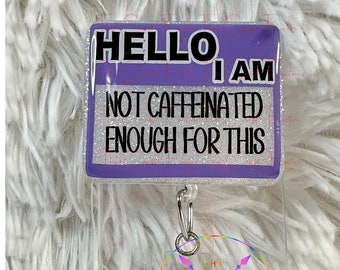 May include: Purple glitter badge holder with the text "HELLO, I AM NOT CAFFEINATED ENOUGH FOR THIS". The badge holder is attached to a clear plastic charm with a rainbow design and the text "THE FIRST"