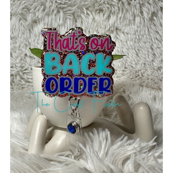 May include: A glittery badge holder with the text "That's on Back Order" in pink, blue, and red lettering. The badge holder has a blue rhinestone charm.