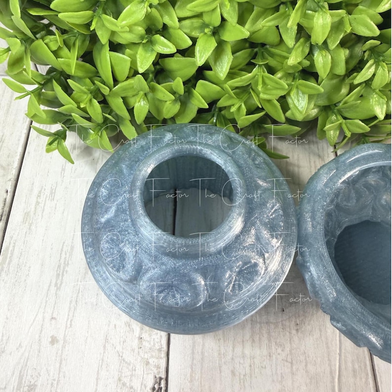 May include: Two light blue decorative resin containers with a circular design and a central opening. The containers have embossed patterns and a glossy finish. The background includes green foliage and a white wooden surface.