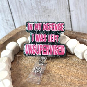 May include: A badge reel with the text "IN MY DEFENSE I WAS LEFT UNSUPERVISED" in pink lettering. The badge reel has a clear plastic holder and a silver-colored metal clip. The text is outlined in a dark green glitter.
