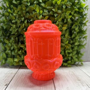 May include: A vibrant orange decorative jar with a lid, featuring intricate embossed patterns. The jar has a cylindrical body with a rounded base and a matching lid. The background includes green foliage and a wooden surface.