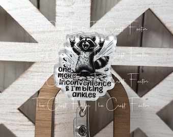 May include: A clear badge reel with a black and white raccoon graphic. The raccoon is making a rock and roll hand gesture. Text on the badge reel reads "one more inconvenience & I'm biting ankles."