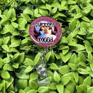 May include: A round, purple badge reel with the text "CURRENT MOOD" and a photo of a dog wearing a blue hood. The badge reel has a clear plastic strap and a metal clip. The background is green.