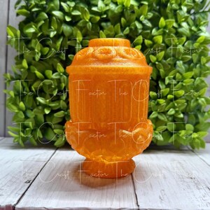 May include: An orange decorative container with a detailed design. The container has a cylindrical body with vertical lines, a decorative base, and a domed lid. The background features green foliage and a white wooden surface.