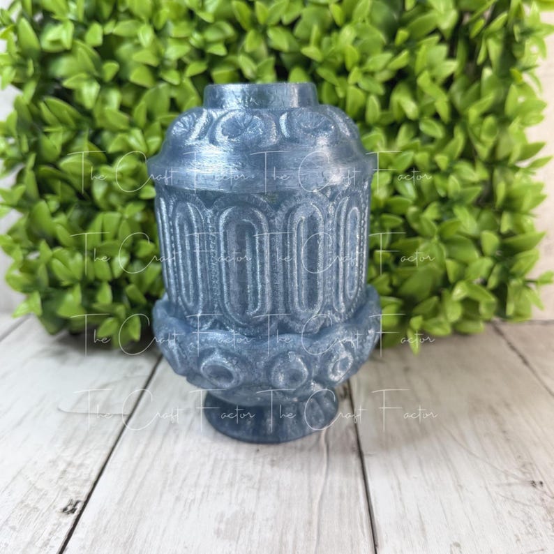 May include: A blue decorative lantern with ornate detailing. The lantern has a cylindrical shape with a base and a top, and is made of a material that appears to have a metallic sheen. The background features green foliage and a wooden surface.