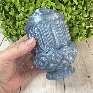 May include: A blue decorative vase with ornate detailing. The vase has a cylindrical shape with vertical grooves and floral accents. The base is round and the top has a flared rim. The Craft Factor is printed on the vase.