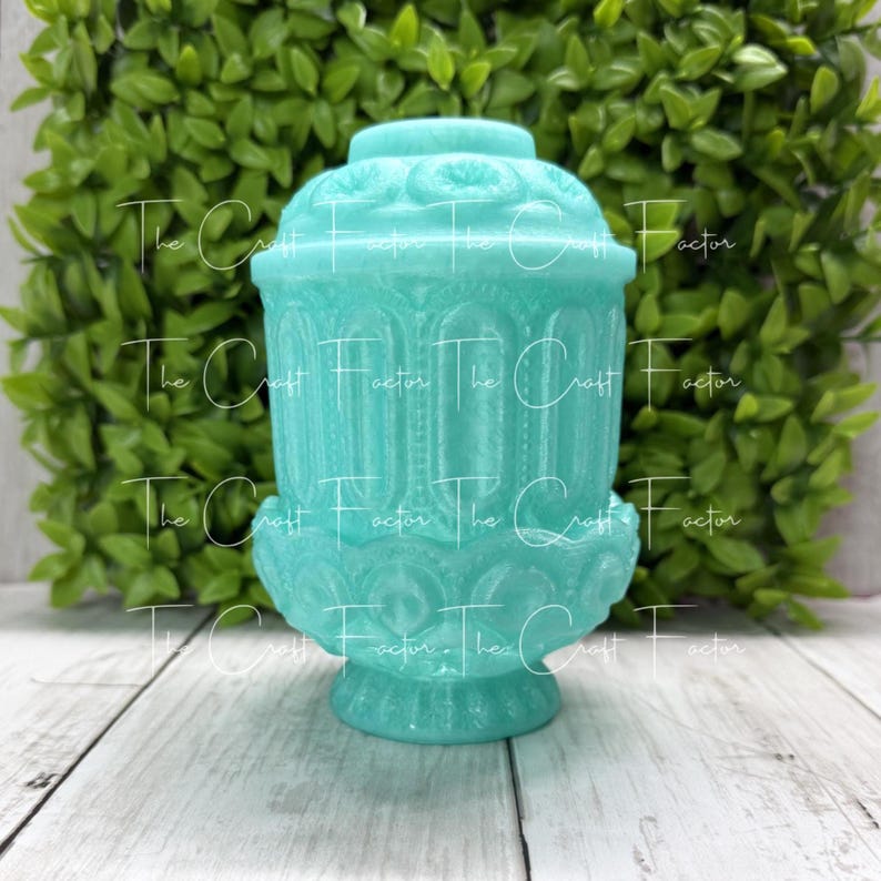 May include: A turquoise decorative jar with a lid, featuring ornate detailing and a pedestal base. The jar has a textured surface and is set against a backdrop of green foliage and a white wooden surface.