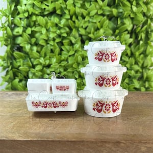 May include: A stack of white ceramic miniature casserole dishes with lids, featuring a red and yellow bird and floral design. The set includes rectangular and round dishes, perfect for a dollhouse or miniature kitchen.
