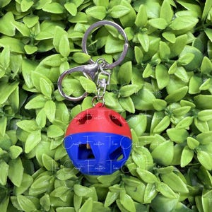 May include: A spherical keychain featuring a red top half and a blue bottom half, both with cut-out geometric shapes. The keychain includes a silver-toned metal split ring and a lobster claw clasp attached to the top of the sphere.