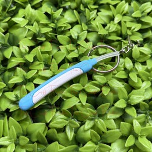 May include: A blue and white keychain with a small, pen-shaped design. The keychain has a silver ring and a clear plastic loop. The background is a bed of green leaves.