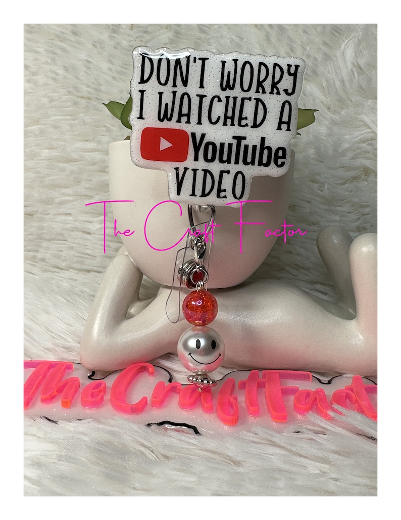 Peut inclure: Porte-badge blanc avec un charme smiley rouge et blanc. Le porte-badge a un fond blanc avec du texte noir qui dit "Ne t'inqui&egrave;te pas, j'ai regard&eacute; une vid&eacute;o YouTube". Le texte est entour&eacute; d'un bouton de lecture YouTube rouge.